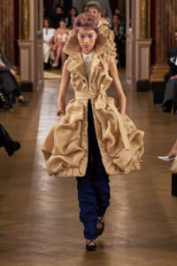 Model walks the runway in a voluminous beige ruffled coat over dark blue pants, during a fashion show.
