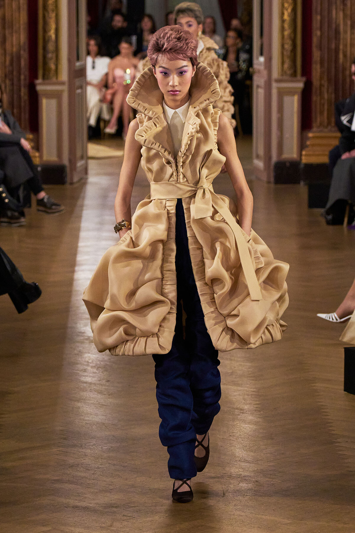 Model walks the runway in a voluminous beige ruffled coat over dark blue pants, during a fashion show.