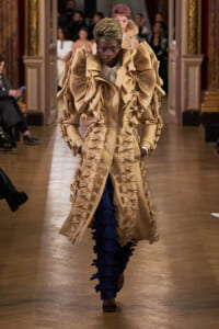 Model walking a fashion runway in a beige, oversized, ruffled coat with bow-like embellishments and blue bow-adorned pants; audience in background.