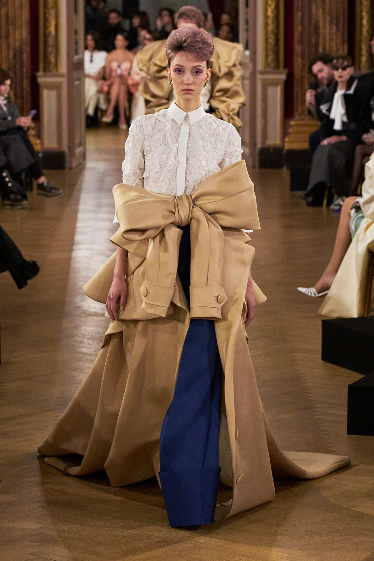 Model walks the runway in a white textured blouse with a large beige cape-like coat tied into a prominent bow at the waist, paired with royal blue wide-legged pants.