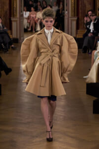 Model walks the runway in a camel-colored oversized trench coat with dramatic puff sleeves, white shirt, and blue shorts visible underneath.