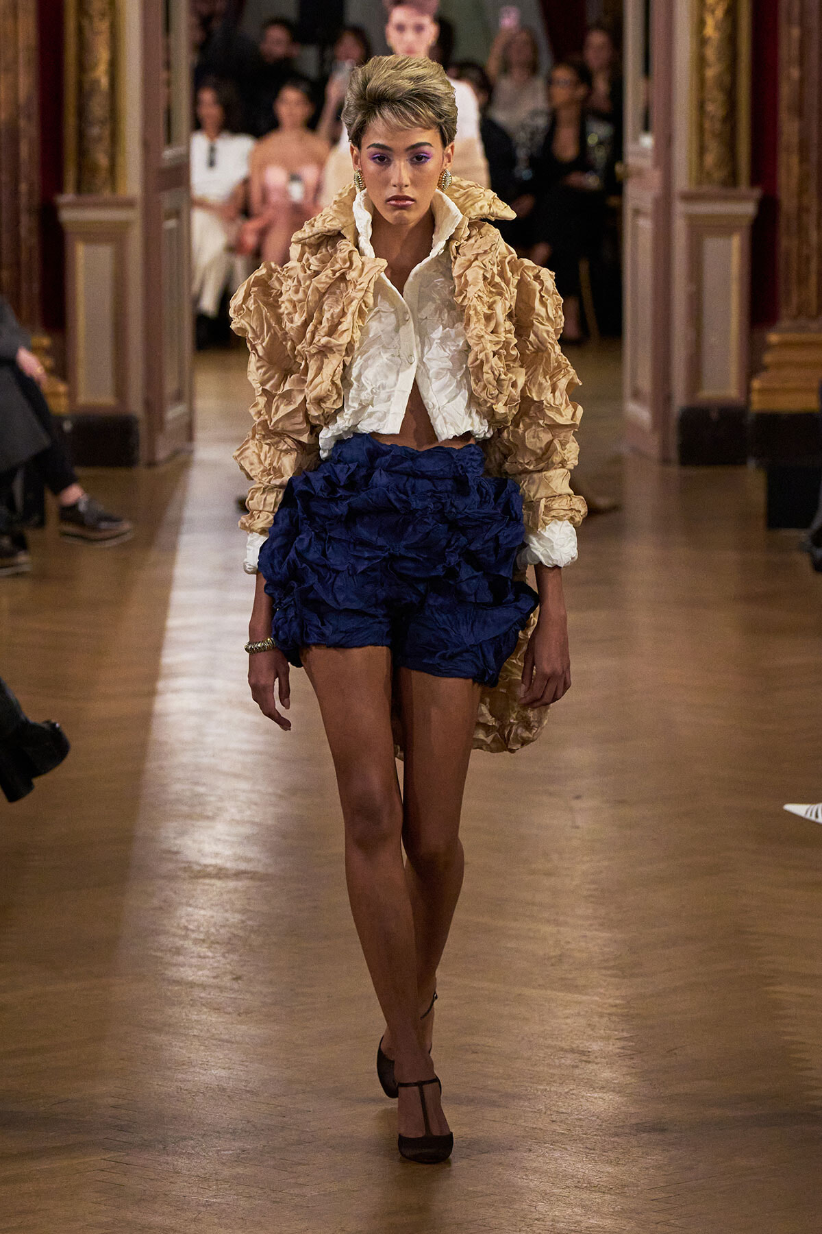 Model walks the runway in a beige, ruffled coat over a white blouse and navy crinkled shorts, heels visible.