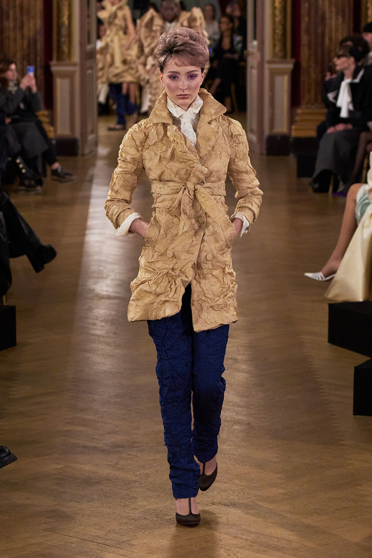 Model walks a fashion runway in a crinkled beige belted coat over a white blouse and blue quilted pants at a show.