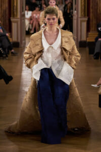 Model walks a runway wearing a tan coat with many ribbon knots over a white blouse and navy wide-leg pants on a catwalk.