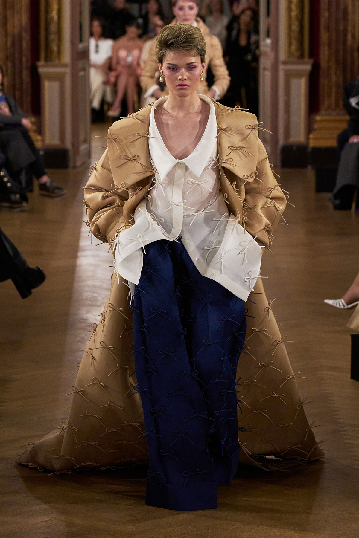 Model walks a runway wearing a tan coat with many ribbon knots over a white blouse and navy wide-leg pants on a catwalk.