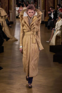 Model walks a runway wearing an oversized beige trench coat with a dramatic high collar and waist belt; blue pants and brown flats visible, audience in background.