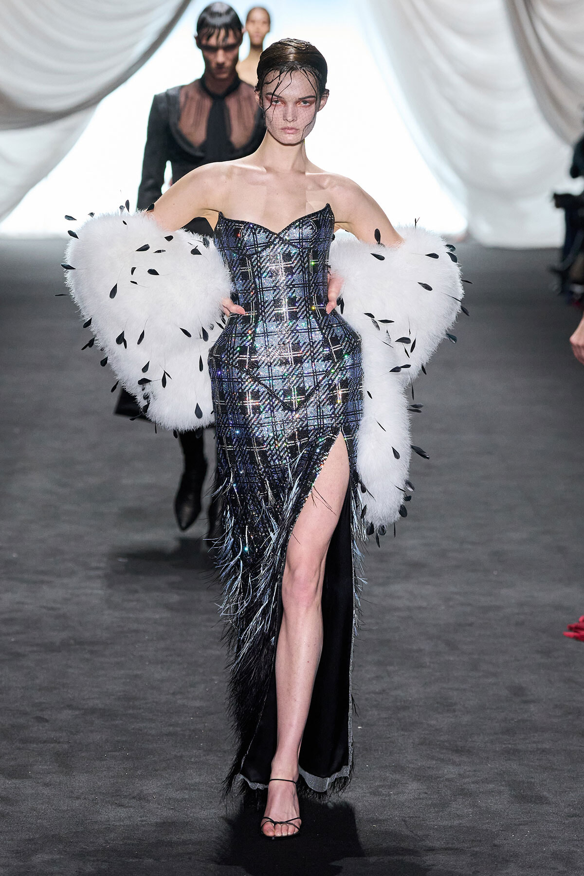 Model on a runway wearing a shimmering plaid corset gown with a high slit and oversized white feather sleeves.