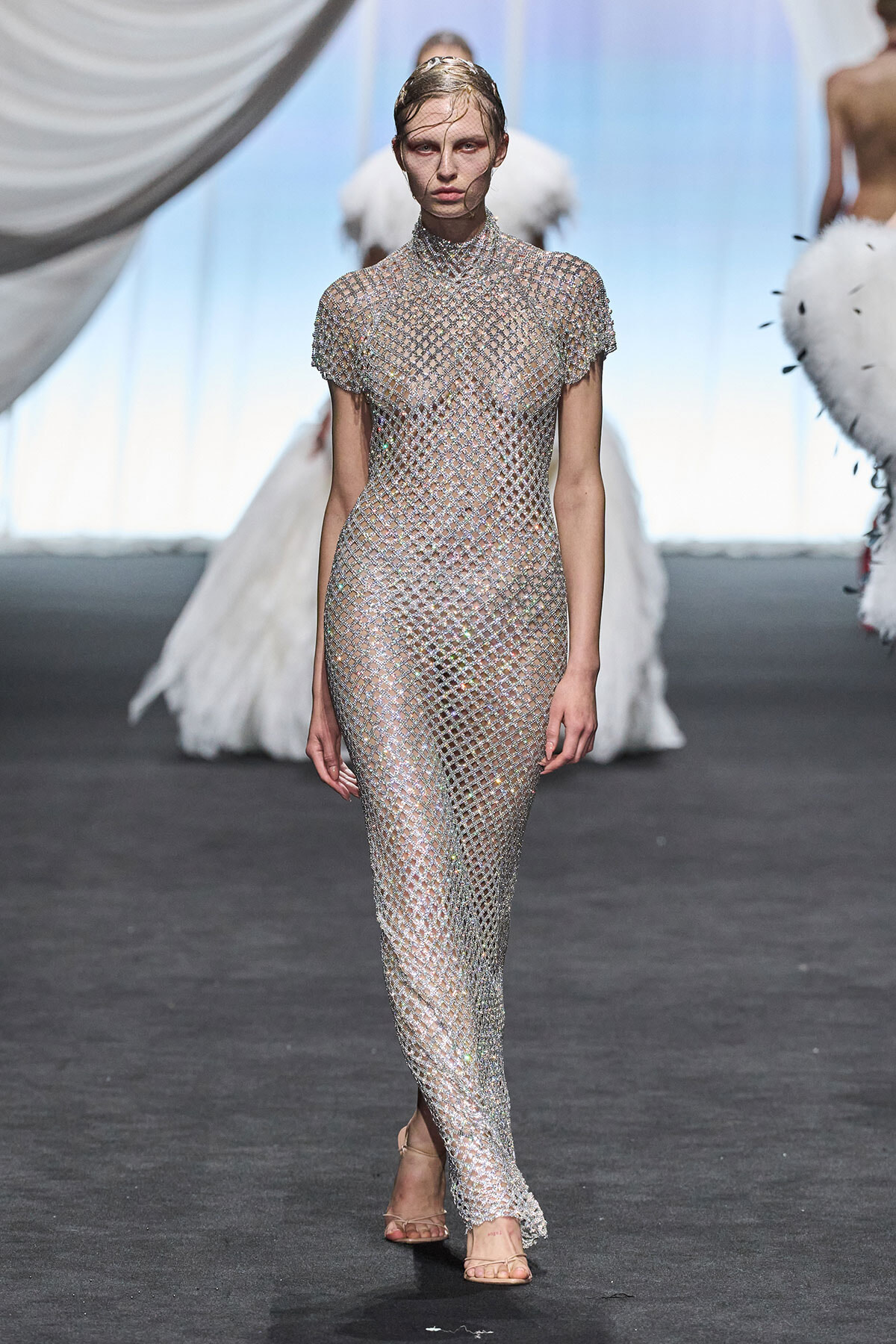Model walks the runway in a full-length, metallic jewelled mesh dress that is sheer and form-fitting, paired with nude sandals.