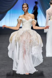 Model on a runway wearing an off-the-shoulder white gown with feathered bodice and voluminous feathered skirt, hands extended as she walks.