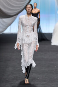 Model walks a runway in a white lace long-sleeve peplum jumpsuit with a sheer face veil and black knee-high boots