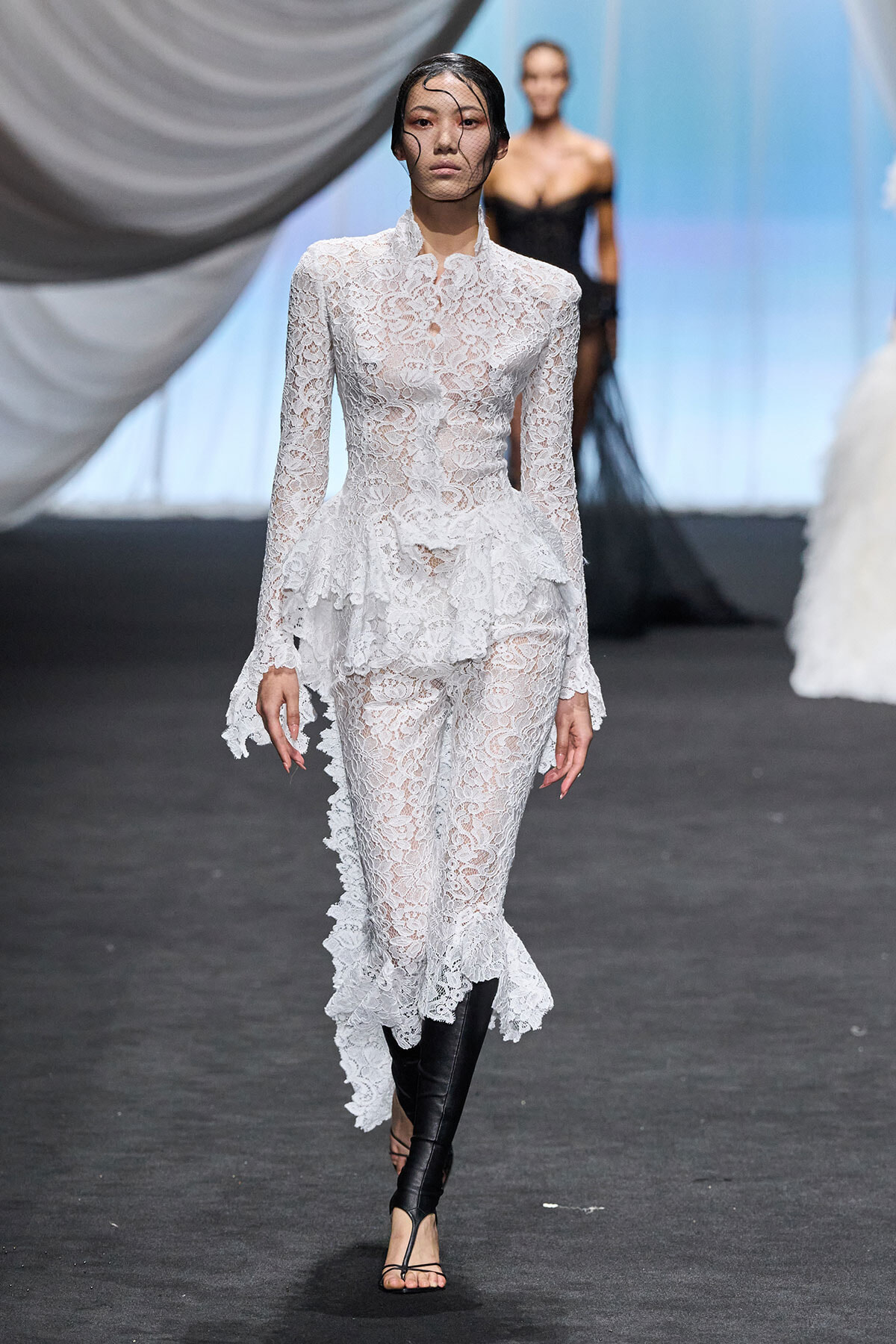 Model walks a runway in a white lace long-sleeve peplum jumpsuit with a sheer face veil and black knee-high boots