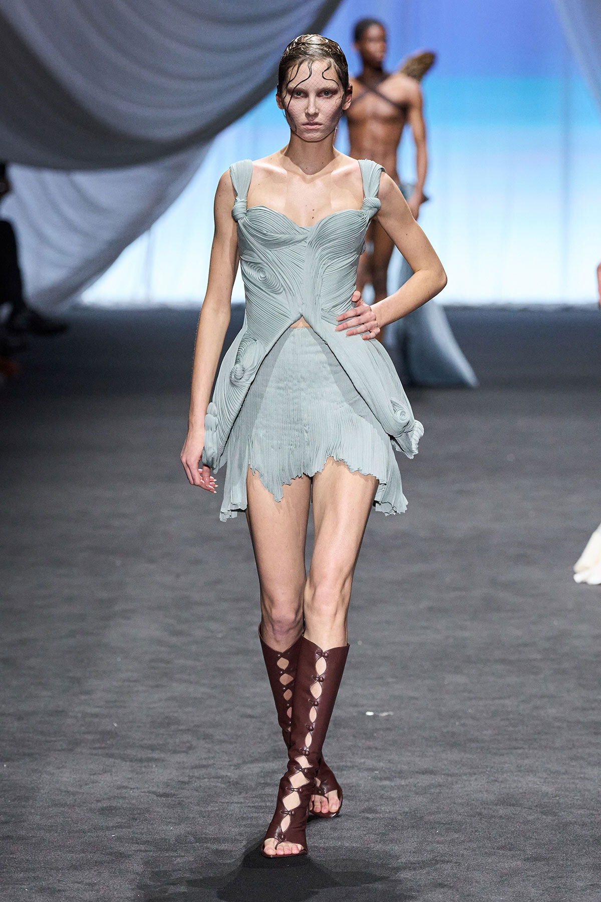 Model on a fashion runway wearing a pale blue sculpted dress with a jagged hem and knee-high brown lace-up boots, blurred background model visible behind.