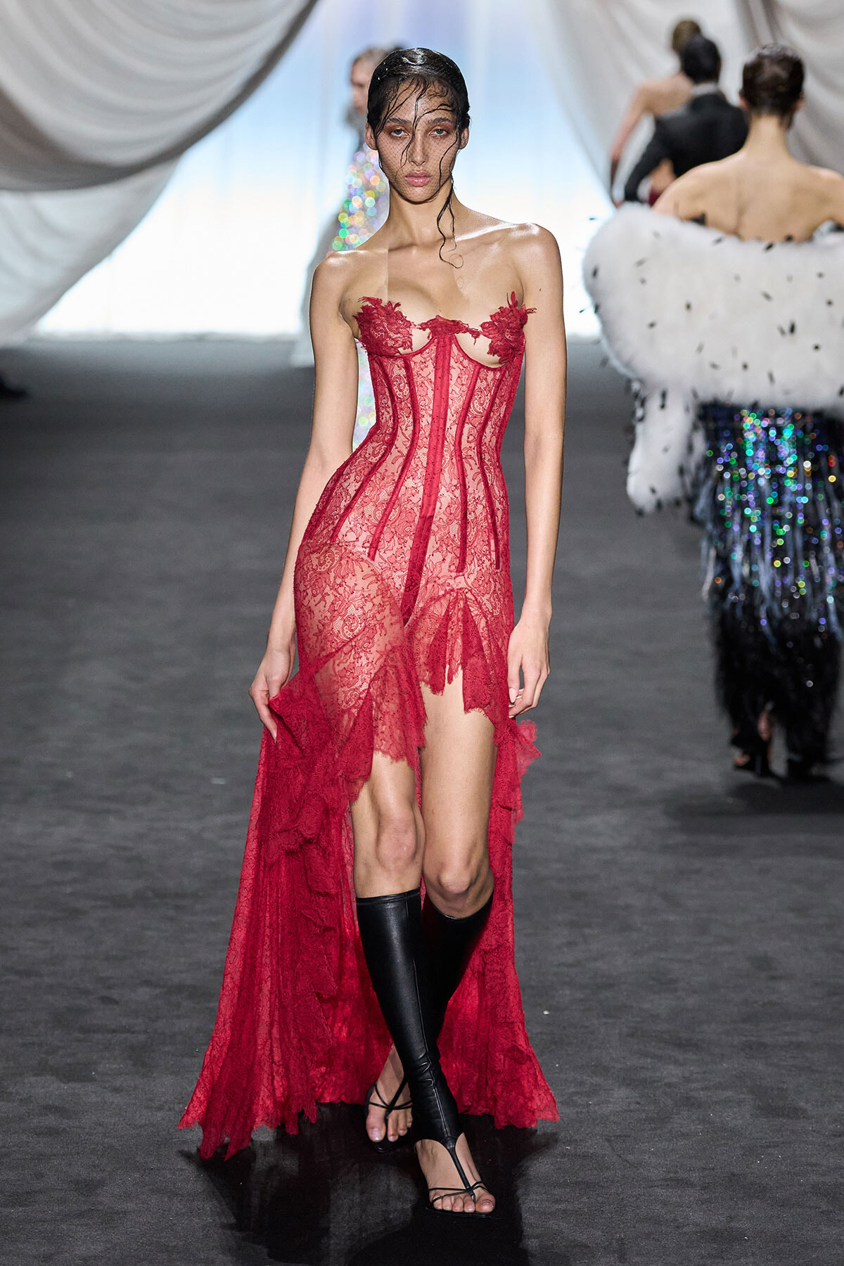 Model walking a runway in a red lace corset gown with sheer panels and a high-low train, paired with black knee-high boots.
