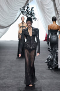 Model on runway wearing a black studded corset with sheer pants and an dramatic jagged headpiece; other models in dark gowns in the background