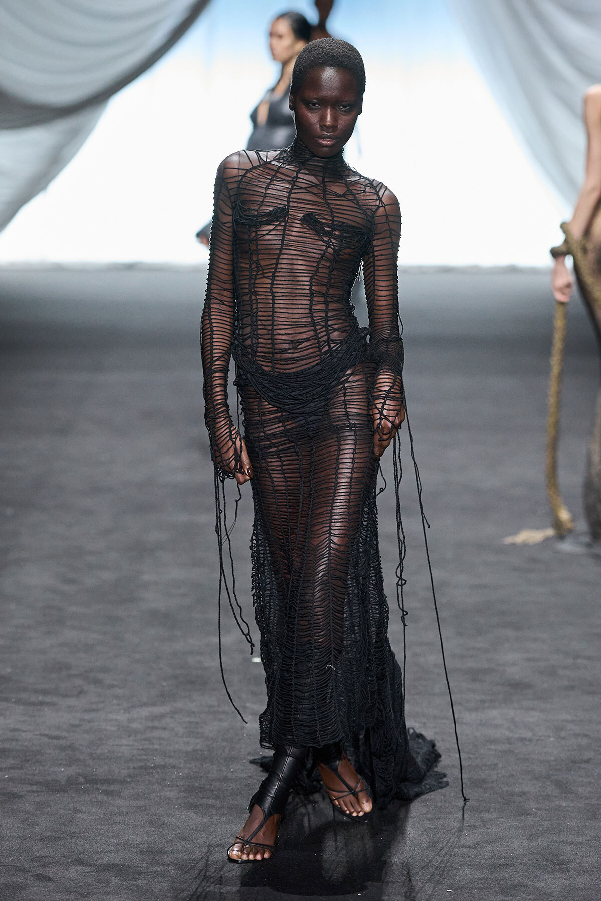 Model walks the runway in a sheer black mesh gown with knotted lines and fringe detailing, wearing flat sandals on a dark catwalk.