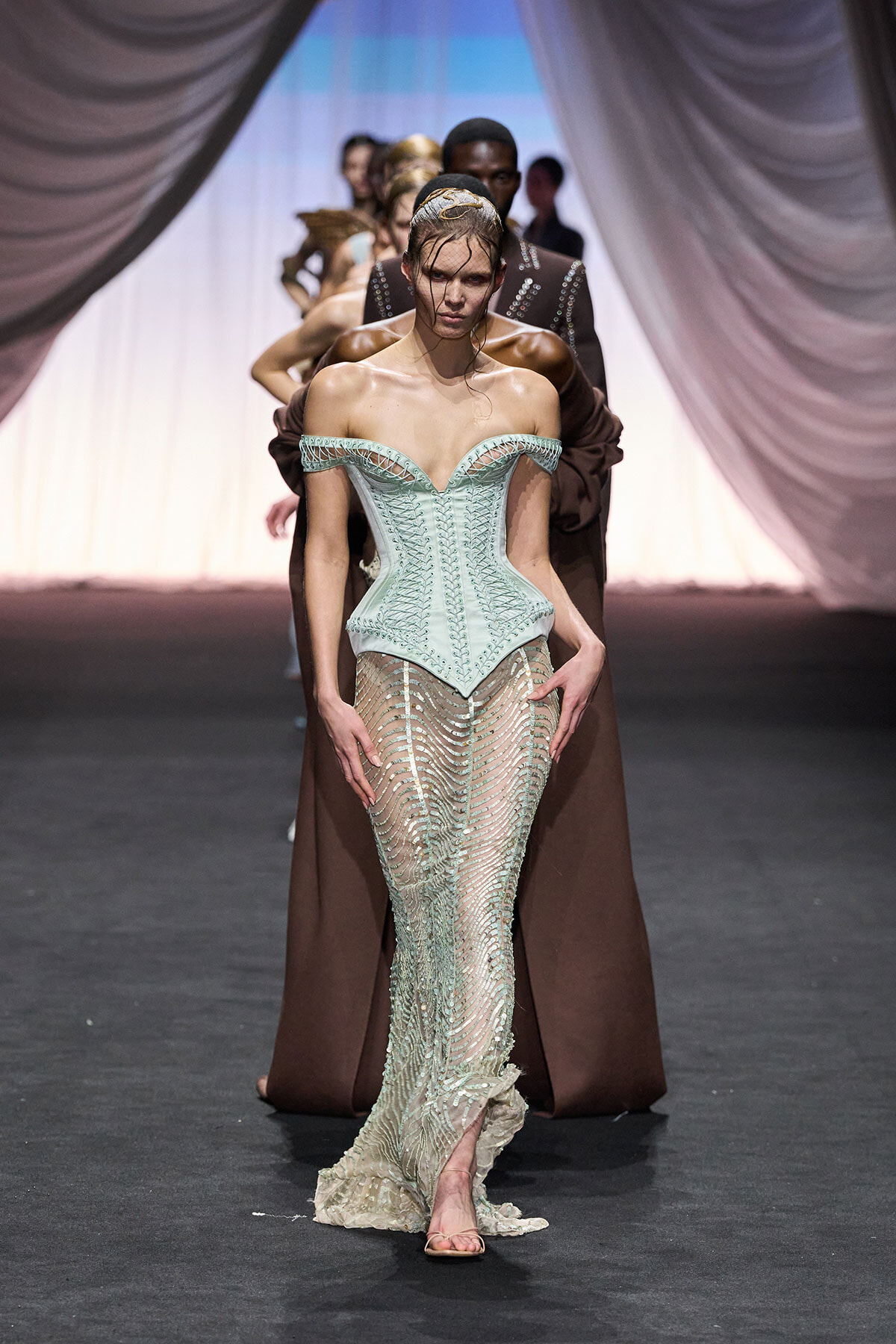 Model walking the runway in a pale mint corset with a sheer, beaded gown; stage drapes and other models in background.