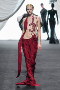 Model walks the runway in a dramatic red gown with a bare midriff and spiral lace details down the front, train trailing behind.