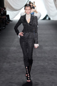 Model walking a runway in a black floral lace pantsuit with a high lace collar, white lace cuffs, and strappy black boots.