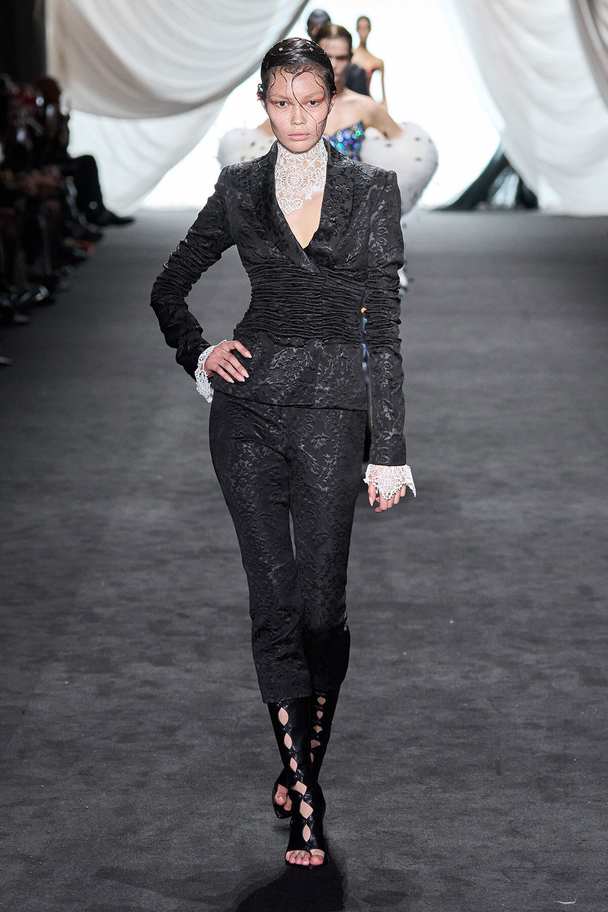 Model walking a runway in a black floral lace pantsuit with a high lace collar, white lace cuffs, and strappy black boots.