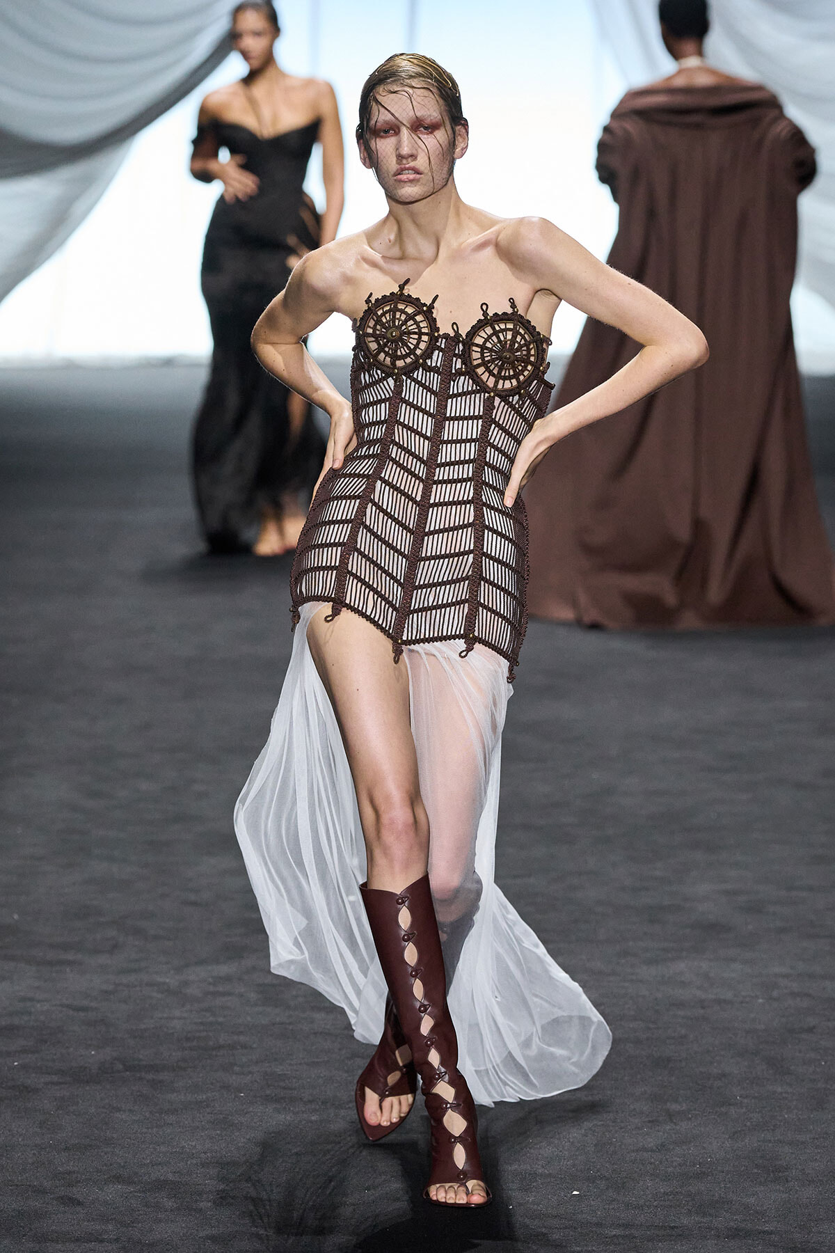 Model walking the runway in a brown crochet corset dress with a sheer white train and knee-high burgundy lace-up boots.