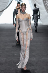 Model walks a runway in a silver corset-style gown with extensive lace-up panels along arms and legs.