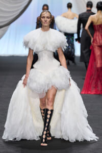Runway model in a dramatic white gown with a voluminous ruffled bodice and corseted waist, paired with black lattice knee-high boots.