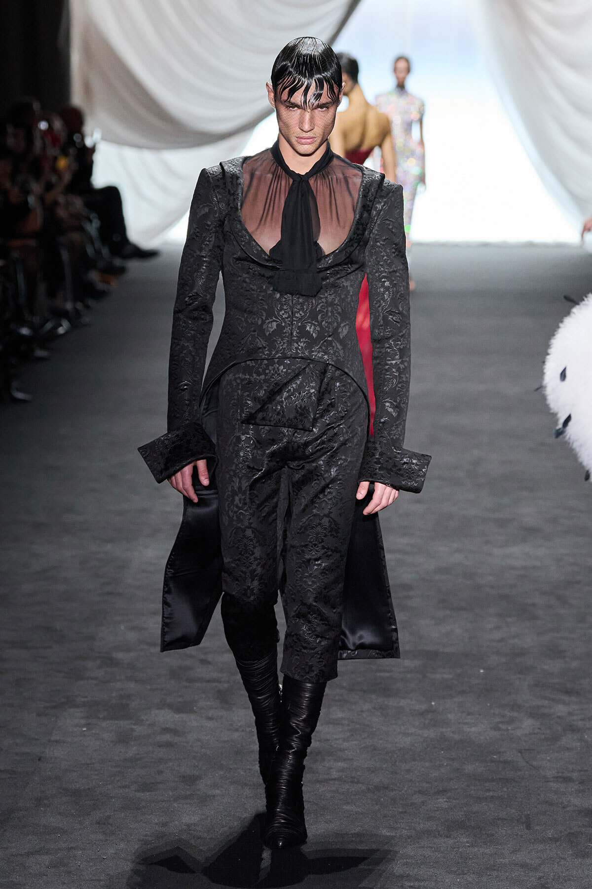 Model walks the runway in a black brocade suit with a sheer blouse and black tie, leather boots, and dramatic fringe hairstyle under bright runway lights.