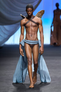 Male runway model walks the catwalk wearing brown briefs, a light blue draped fabric around his waist, and feathered wings accessory in a fashion show.