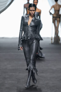 Model walking a fashion show in a charcoal crocodile-textured suit with a peplum and matching knee-high boots.