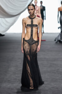 Model on the runway wearing a sheer black lace gown with a bold cross motif across the torso and a high, lace-neck collar, standing on the catwalk.