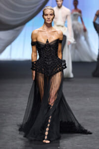 Model on a runway wearing a black off-shoulder lattice corset gown with a sheer, pleated skirt and lace-up sandals in a fashion show setting.
