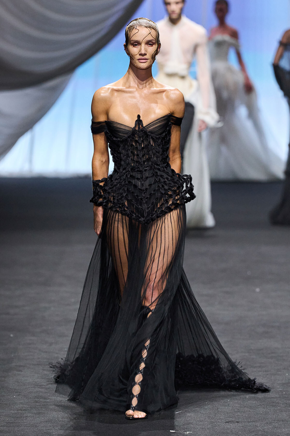 Model on a runway wearing a black off-shoulder lattice corset gown with a sheer, pleated skirt and lace-up sandals in a fashion show setting.