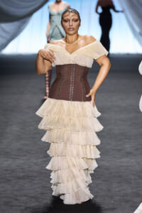 Model walking a runway in an off-shoulder cream top, brown lace-up corset, and a multi-tiered cream ruffle skirt with a sheer face veil.