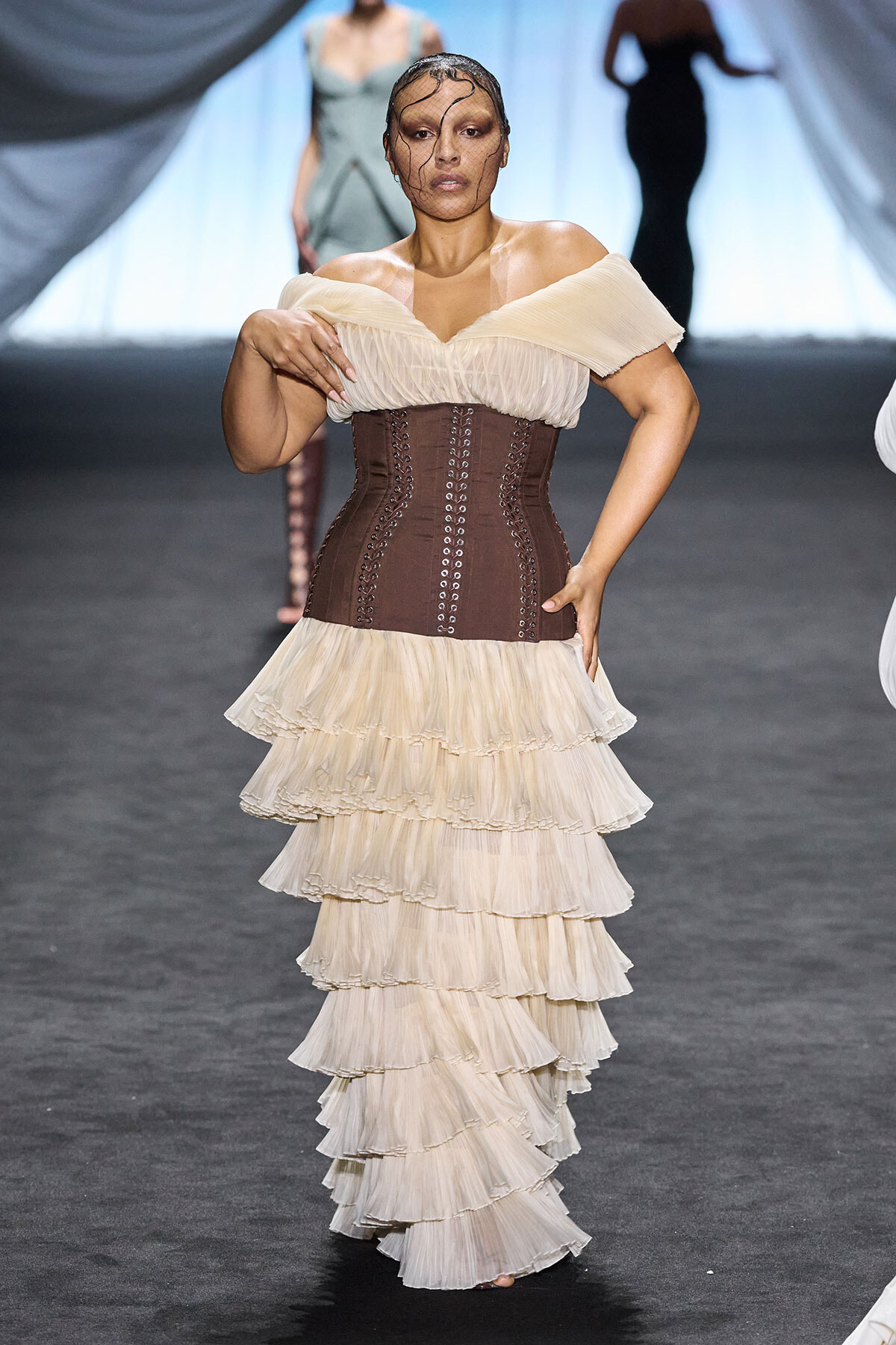 Model walking a runway in an off-shoulder cream top, brown lace-up corset, and a multi-tiered cream ruffle skirt with a sheer face veil.