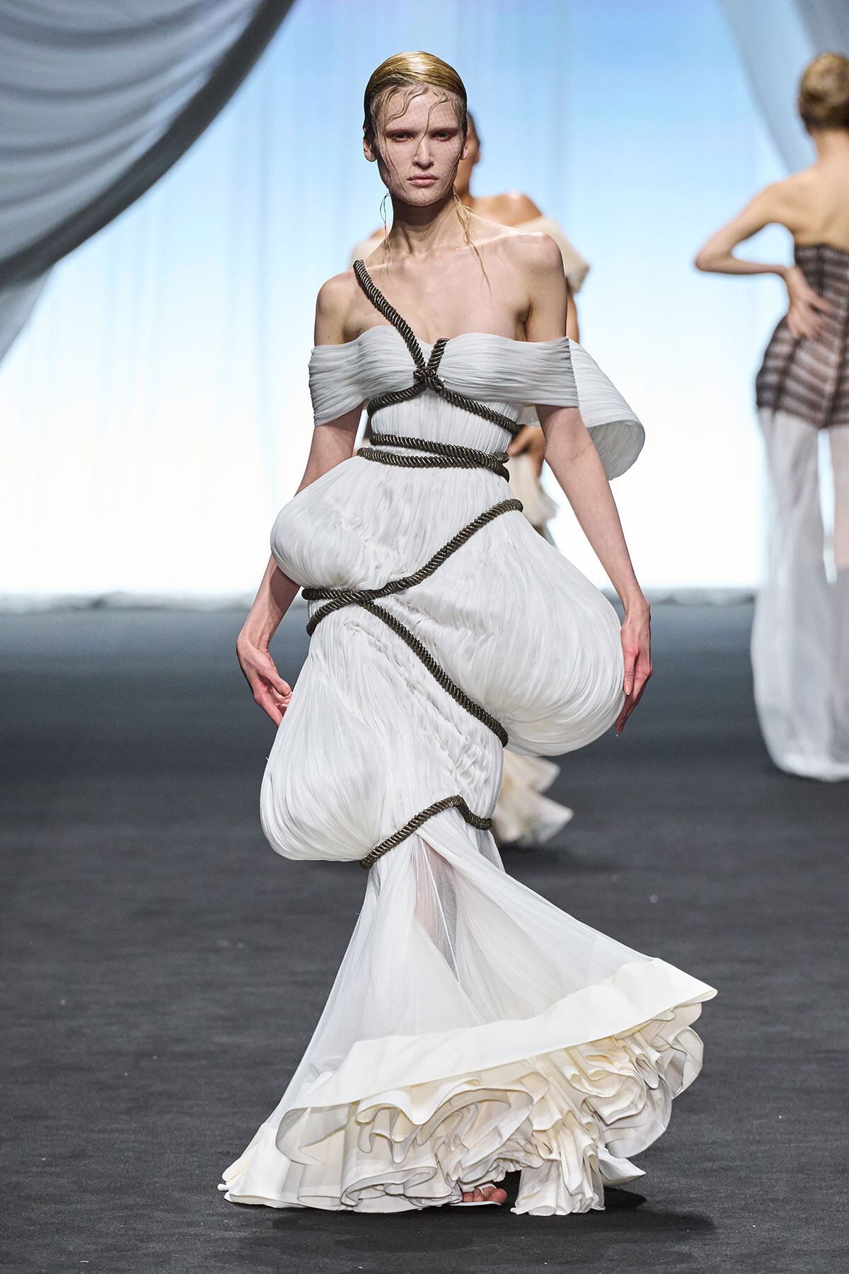 Model walks the runway in an off-shoulder white gown with a voluminous ruched skirt wrapped in dark rope.