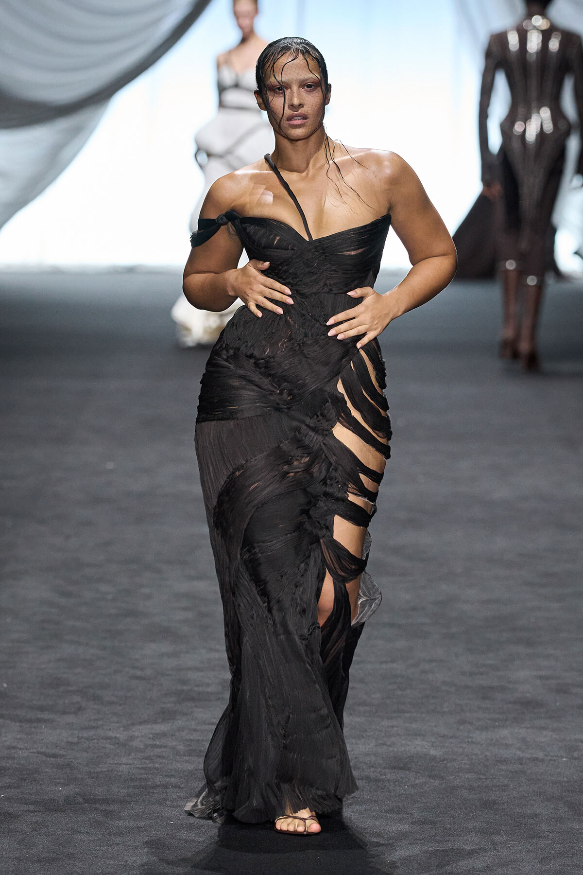 Runway model walking in a black off-shoulder gown with torn, diagonal panels and sheer accents.