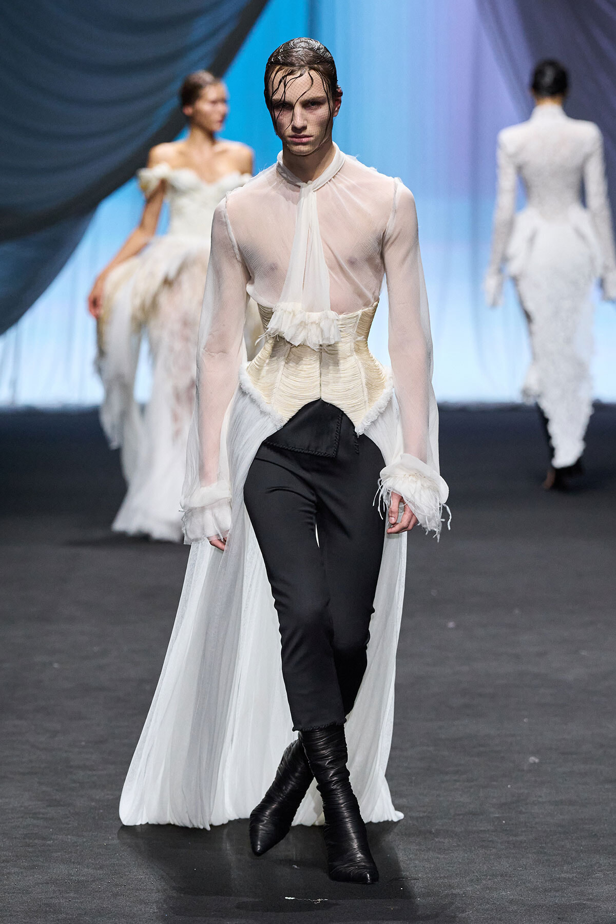 Model walks the runway in a sheer ivory blouse with a sculpted corset over black pants and tall black boots, cape trailing behind.