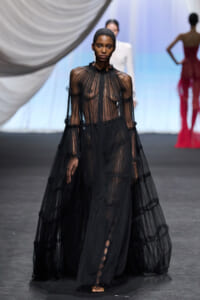 Model walking the runway in a sheer, black, floor-length gown with tiered layers and long sleeves.