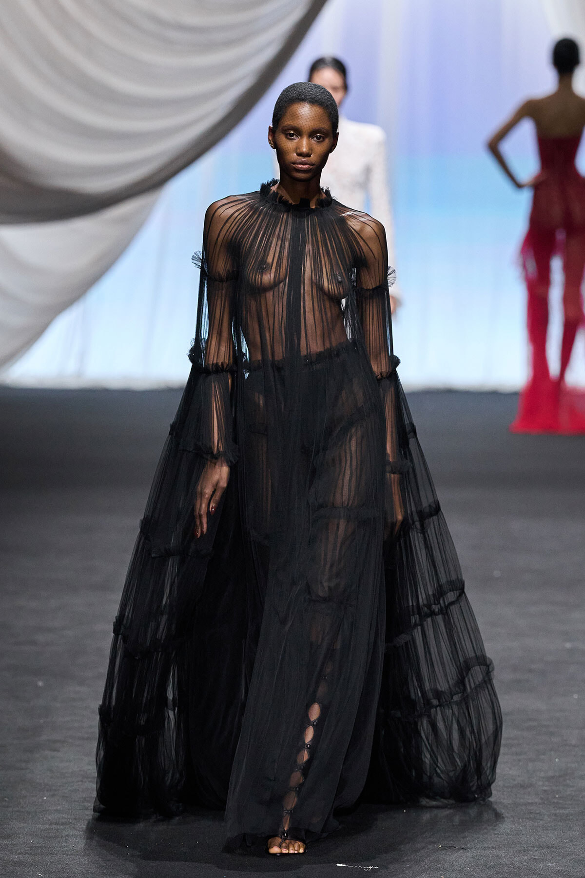 Model walking the runway in a sheer, black, floor-length gown with tiered layers and long sleeves.
