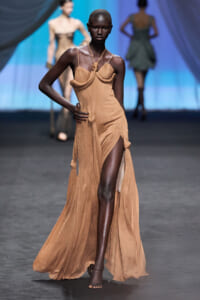 Model walking a runway in a tan, pleated gown with a high slit and sculpted draped bodice.