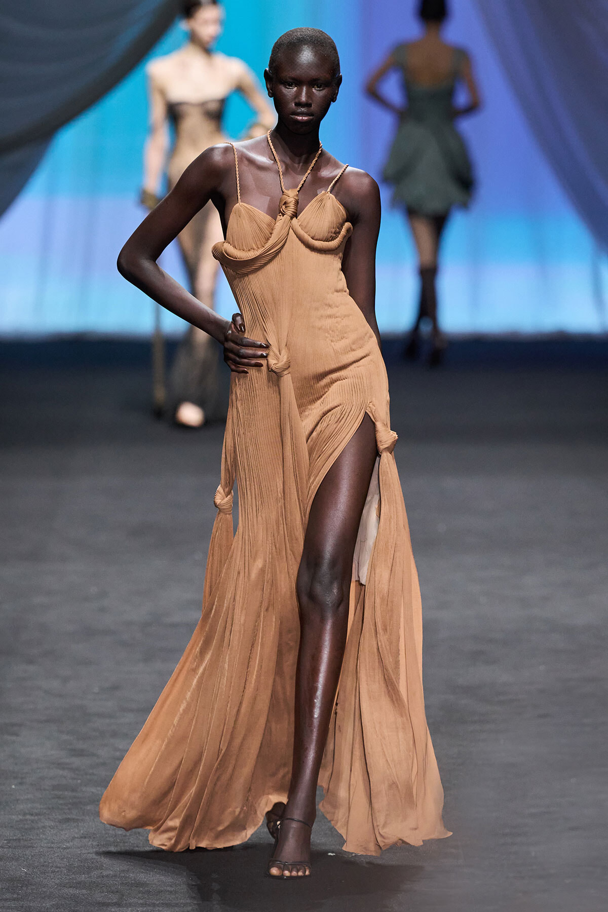 Model walking a runway in a tan, pleated gown with a high slit and sculpted draped bodice.