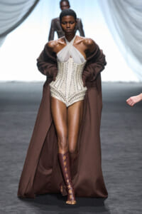 Model on a runway wearing a white cross-front corset bodysuit with a long brown cape and matching knee-high lace-up boots.