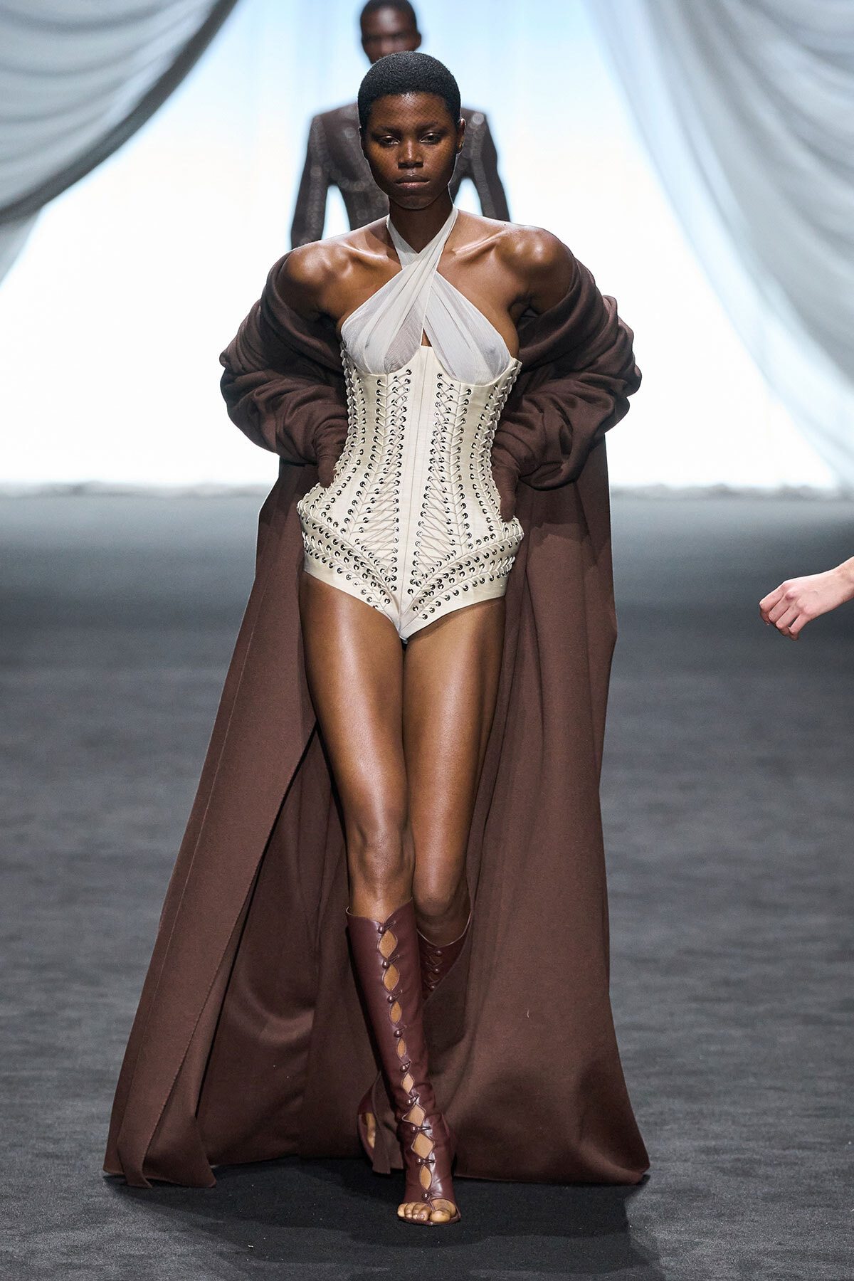 Model on a runway wearing a white cross-front corset bodysuit with a long brown cape and matching knee-high lace-up boots.