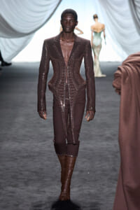 Male model walks the runway in a dark brown, heavily laced bodysuit with metal eyelets and knee-high boots.
