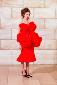 Model in a vivid red sculptural dress with oversized bow-like ruffles, off-shoulder, standing against a pale stone wall.