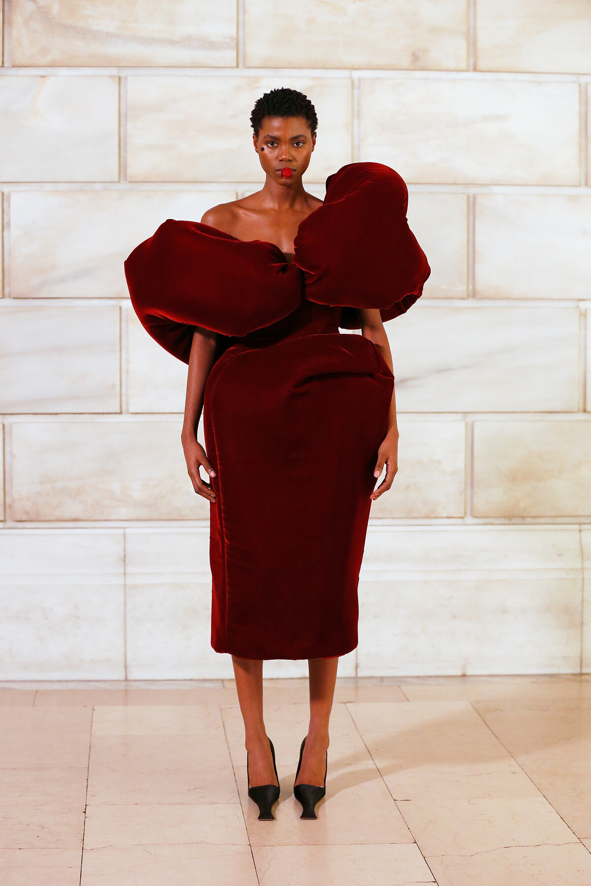 Model wearing a dramatic red velvet dress with oversized bow shoulders against a light stone wall.