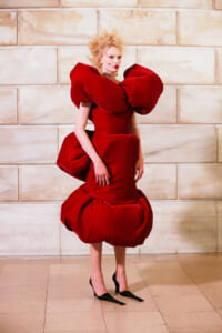 Model wearing a dramatic red sculptural dress with oversized ruffled shoulders and a voluminous bubble skirt against a beige stone wall.