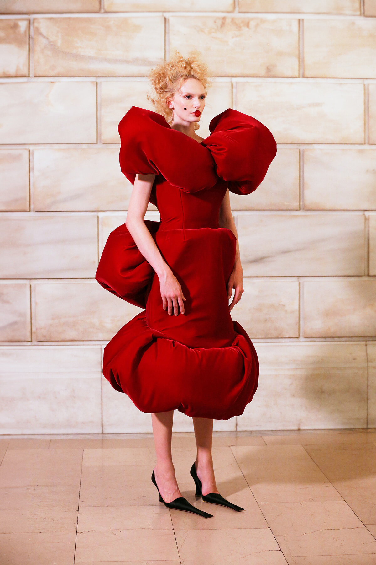 Model wearing a dramatic red sculptural dress with oversized ruffled shoulders and a voluminous bubble skirt against a beige stone wall.