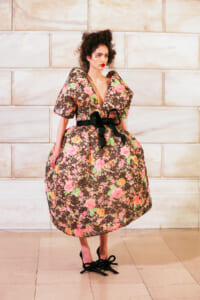 Model wearing a voluminous floral dress with a black sash, standing against a light stone wall.