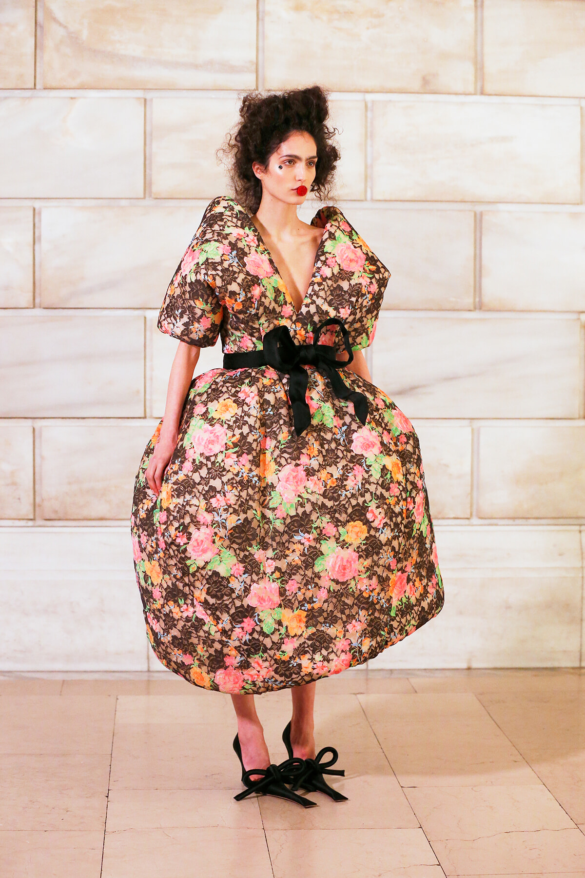 Model wearing a voluminous floral dress with a black sash, standing against a light stone wall.